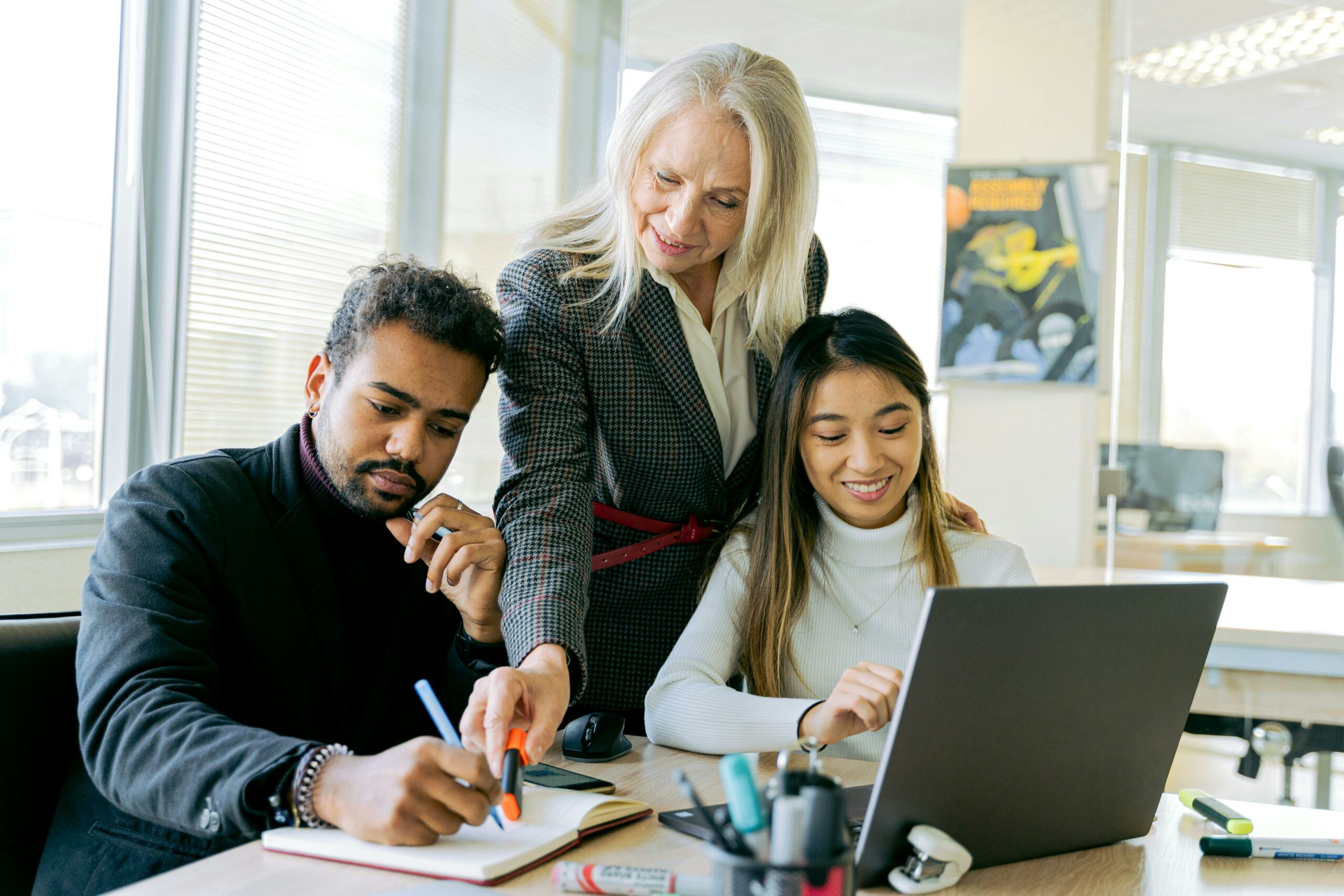 Trois collègues travaillant ensemble dans un bureau, une femme plus âgée accompagne deux jeunes adultes sur un projet.