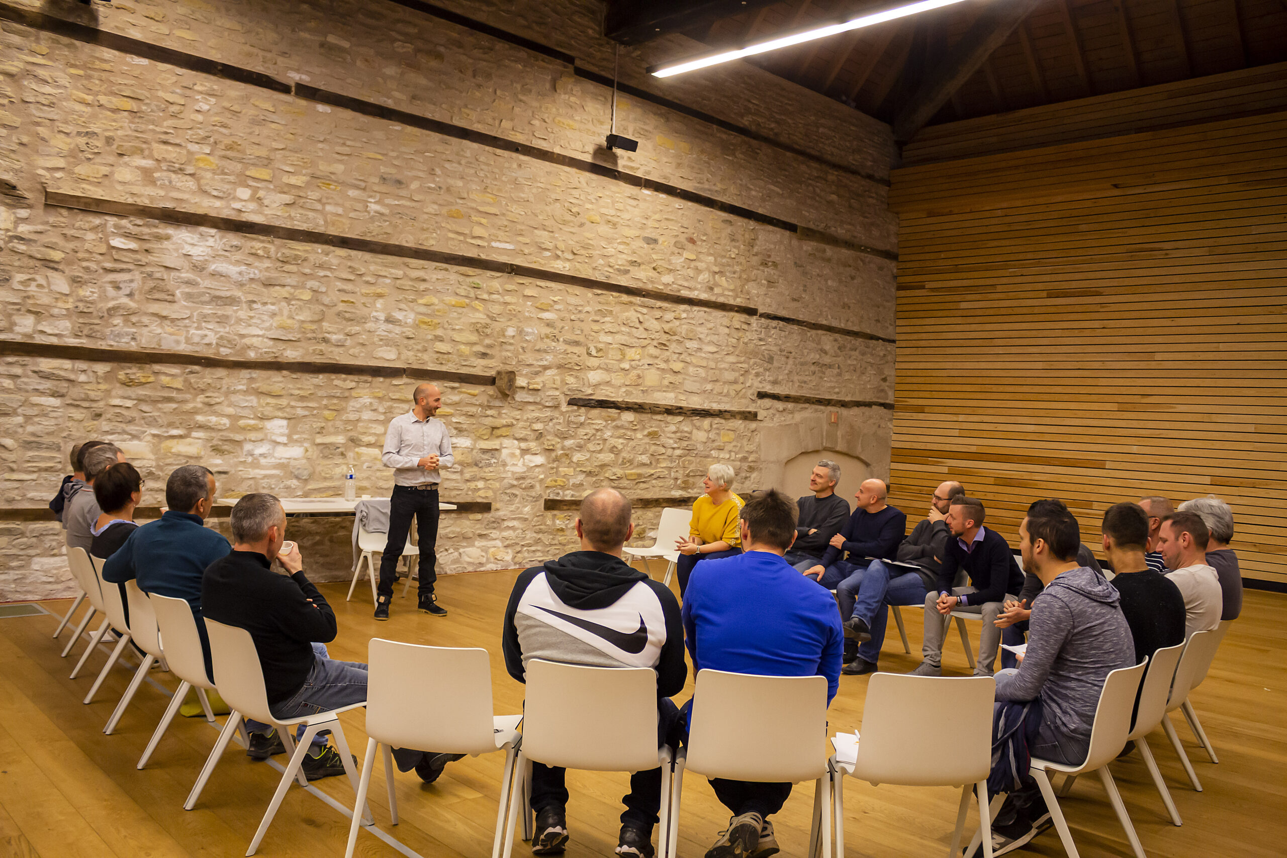 Un intervenant anime un atelier de bien-être devant un groupe de collaborateurs assis en cercle dans une salle en pierre.